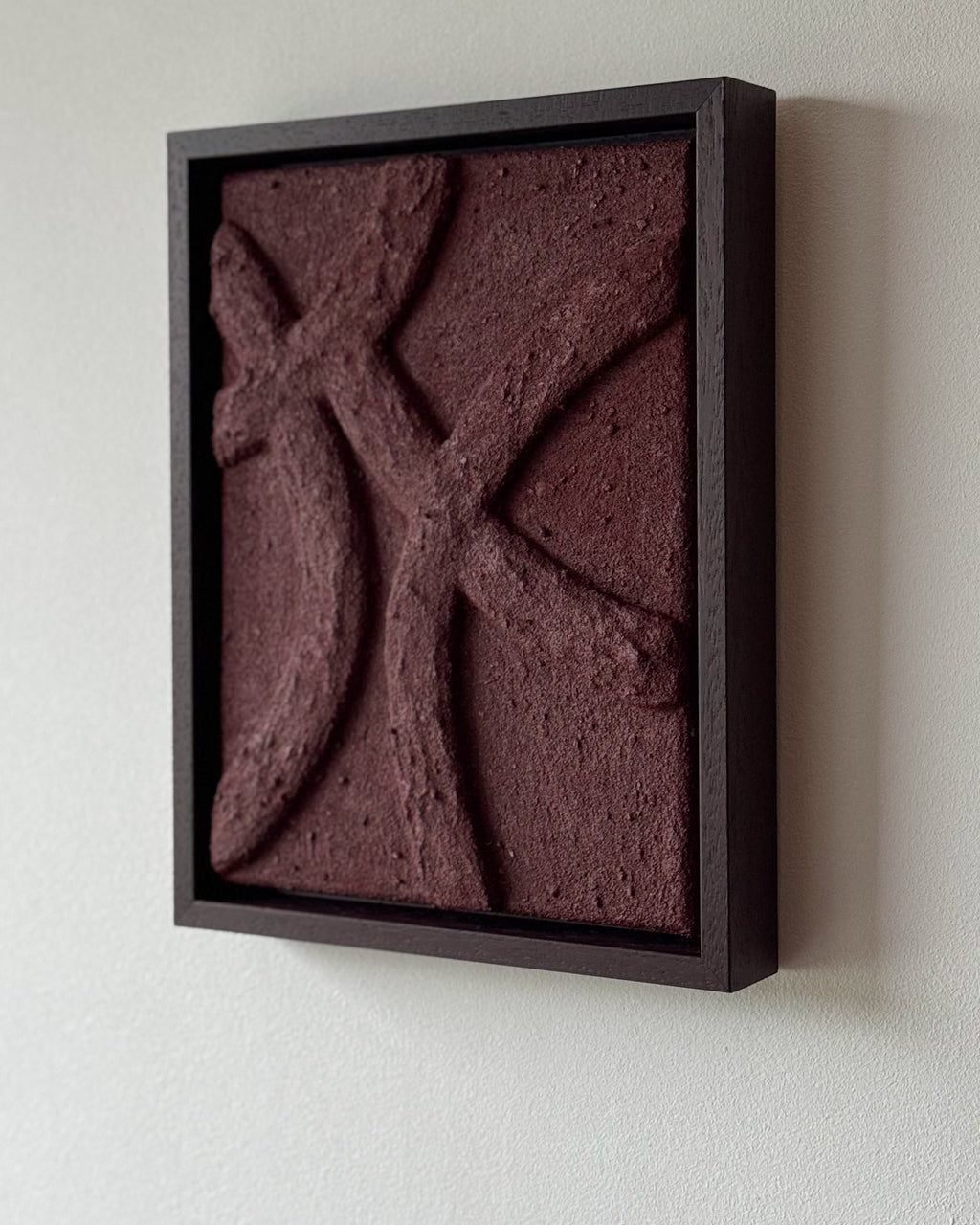 Een handgemaakt abstract reliëf kunstwerk in bordeauxrood met houten lijst en organische lijnen en structuur aan de muur.