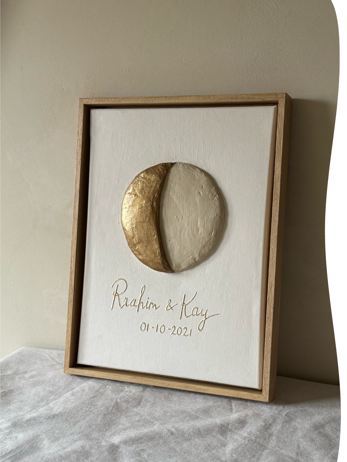 Maanfase reliëfkunstwerk van de trouwdag van Rhahim en Kay in neutrale kleuren tegen muur.